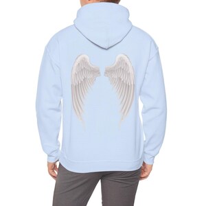 May include: A light blue hoodie featuring a detailed illustration of white angel wings on the back. The wings have a layered feather design with subtle shading, creating a realistic appearance. The hoodie has a hood and a relaxed fit.