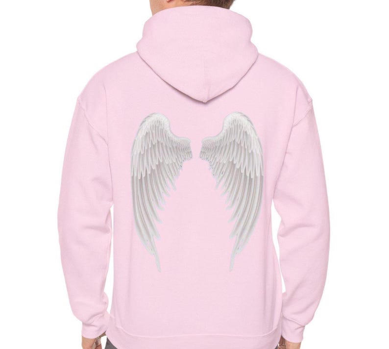 May include: A light pink hoodie with a hood and white angel wings printed on the back. The wings are detailed with individual feathers, creating a realistic appearance. The hoodie is a casual garment.