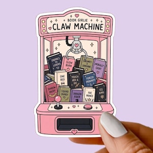 May include: A pink claw machine sticker featuring the text BOOK GIRLIE CLAW MACHINE at the top. The machine is filled with various books labeled with tropes like Enemies to Lovers, Slow Burn, One Bed, and Forced Proximity, with a metal claw above.