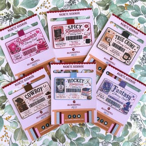 May include: Six rectangular magnetic bookmarks on white backing cards, each featuring a unique library card design with themes like Romance, Spicy Romance, True Crime, Cowboy Romance, Hockey Romance, and Fantasy, all labeled Bloomspell Press.
