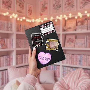 May include: A black e-reader decorated with four stickers: a white rectangular warning label, a black book sticker with the text 'HE'S A RED FLAG I'M COLORBLIND', a crime scene themed sticker, and a pink heart sticker with the text 'They be fucking in this book'.