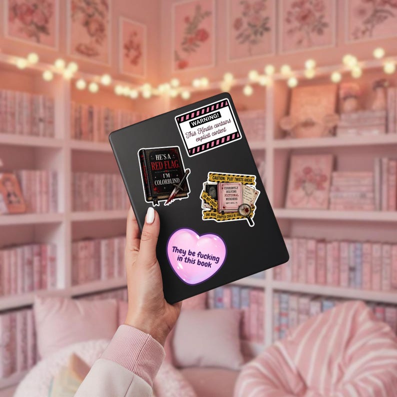May include: A black e-reader decorated with four stickers: a white rectangular warning label, a black book sticker with the text 'HE'S A RED FLAG I'M COLORBLIND', a crime scene themed sticker, and a pink heart sticker with the text 'They be fucking in this book'.
