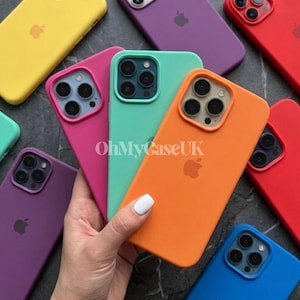 Könnte beinhalten: Eine Auswahl an bunten Handyhüllen in Gelb, Lila, Rot, Orange, Pink, Grün und Blau. Jede Hülle hat einen Kameraausschnitt und das Apple-Logo. Die Hüllen sind auf einer dunklen Oberfläche angeordnet.