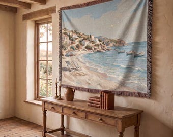 May include: A woven wall tapestry featuring a coastal landscape with a sandy beach, blue ocean water, small boats, and hillside houses. The rectangular textile has a fringed border and displays a soft color palette of light blue, beige, and white.