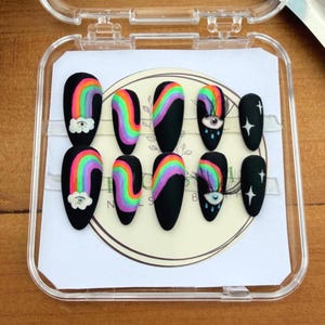 以下が含まれることがあります： カラフルな虹のデザイン、雲のアクセント、目のモチーフが施された、黒いアクリルネイル10本セット。 ネイルは透明なプラスチックケースに収められており、鮮やかなネイルアートが際立っています。