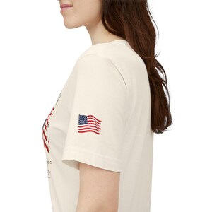 May include: A cream-colored short-sleeve t-shirt featuring a small American flag graphic printed on the sleeve. The shirt has a crew neckline and a partial graphic design visible on the front chest area, showcasing red and blue ink details.