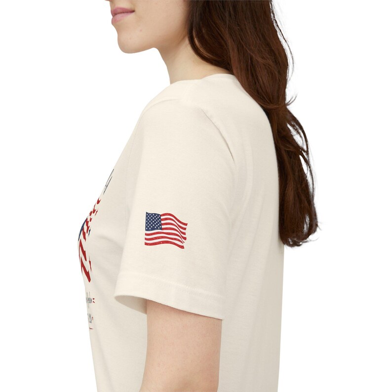 May include: A cream-colored short-sleeve t-shirt featuring a small American flag graphic printed on the sleeve. The shirt has a crew neckline and a partial graphic design visible on the front chest area, showcasing red and blue ink details.