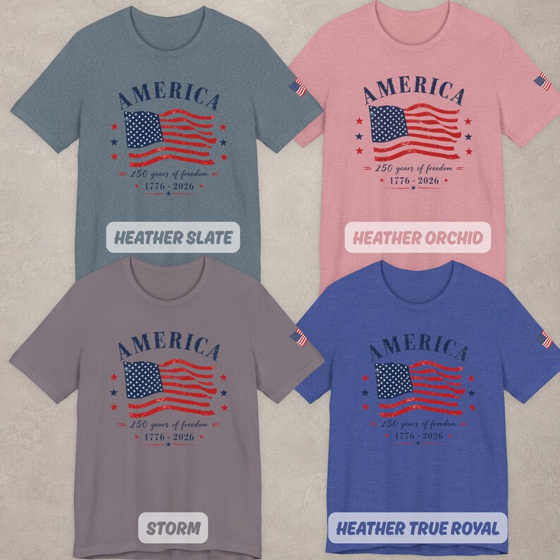 May include: Four short-sleeve t-shirts in heather slate, heather orchid, storm, and heather true royal colors. Each shirt features a graphic of an American flag with the text AMERICA, 250 years of freedom, and 1776-2026, plus a small flag on the sleeve.