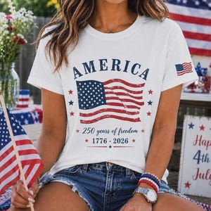 May include: A white short-sleeve t-shirt featuring the word AMERICA in blue, a graphic of a waving American flag, and the text 250 years of freedom 1776-2026. A small American flag graphic is printed on the right sleeve of the garment.