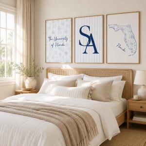 May include: Three framed wall art prints hanging above a bed. The left print features a blue floral pattern with the text The University of Florida. The centre print displays a large blue SA monogram on a striped background. The right print shows a blue floral map of Florida with the text Florida.