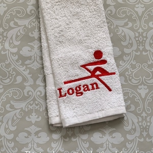 May include: White hand towel with red embroidery of a person rowing a boat and the name "Logan".