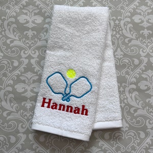 May include: White towel with blue and yellow embroidered pickleball paddles and the name "Hannah" in red.