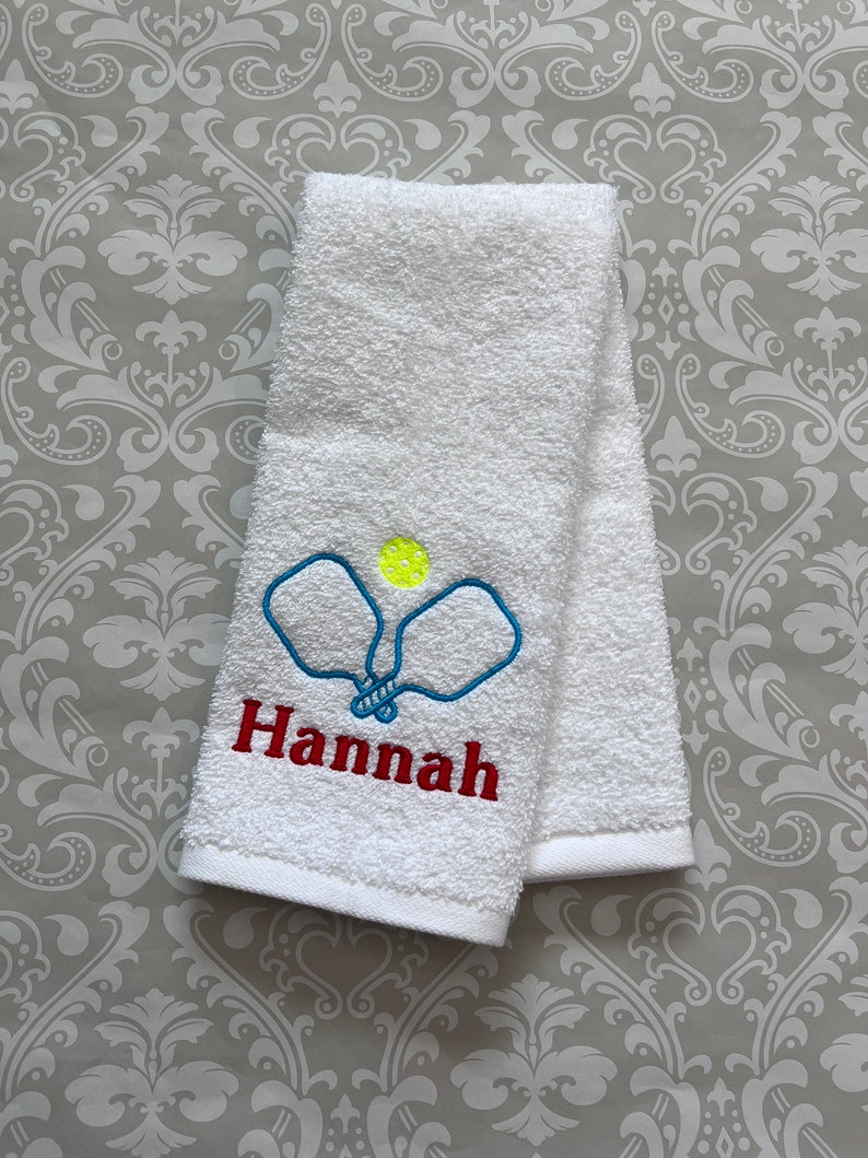 May include: White towel with blue and yellow embroidered pickleball paddles and the name "Hannah" in red.