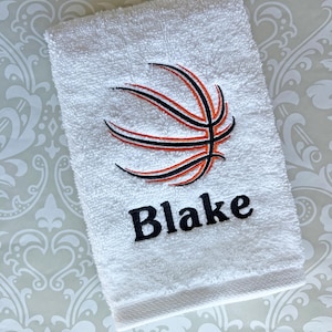 May include: White hand towel embroidered with a basketball design in black and orange. The name "Blake" is embroidered in black block letters below the basketball graphic. The towel has a textured surface and a decorative border.