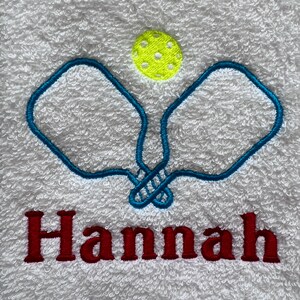 May include: White towel with blue embroidered pickleball paddles and a yellow pickleball. The name "Hannah" is embroidered in red below the paddles.