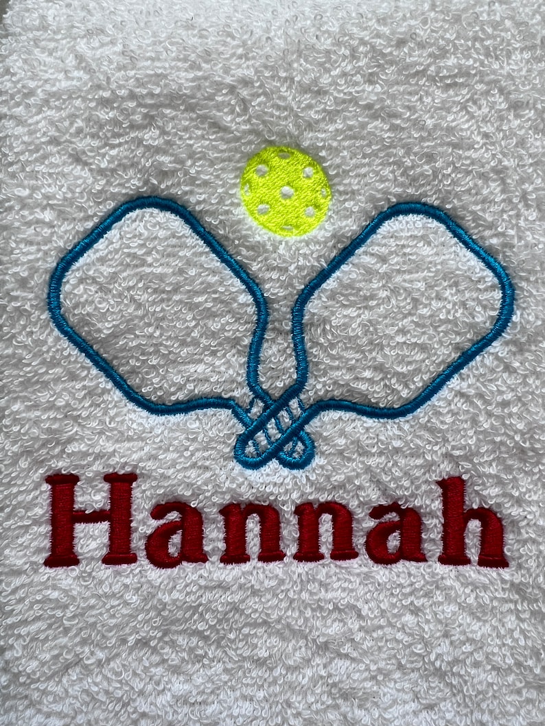 May include: White towel with blue embroidered pickleball paddles and a yellow pickleball. The name "Hannah" is embroidered in red below the paddles.