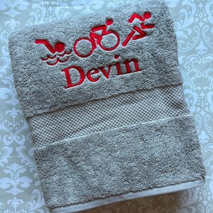 May include: A light gray towel with red embroidered triathlon symbols and the name "Devin." The towel features a swimming, cycling, and running graphic. The text at the bottom of the image reads: "This light gray towel is stitched in red."