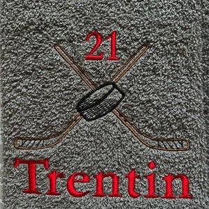 May include: Gray towel with red embroidered text "Trentin" and the number "21". Two crossed hockey sticks with a black puck are embroidered above the text.