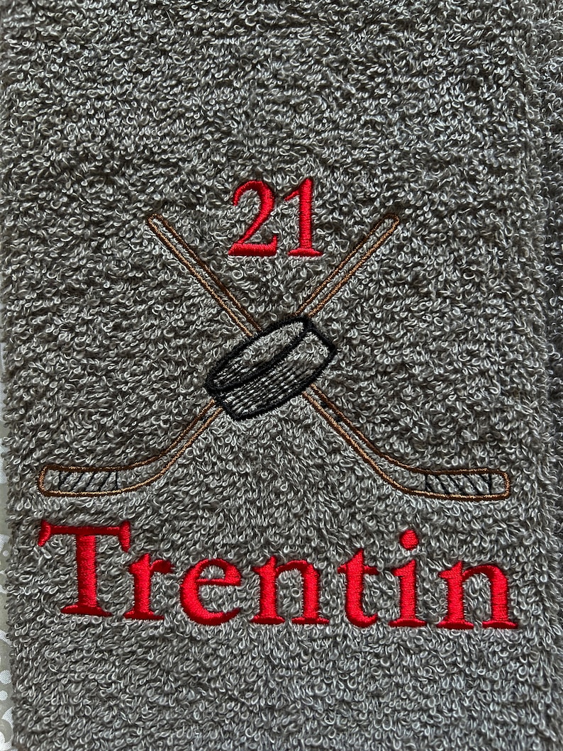 May include: Gray towel with red embroidered text "Trentin" and the number "21". Two crossed hockey sticks with a black puck are embroidered above the text.