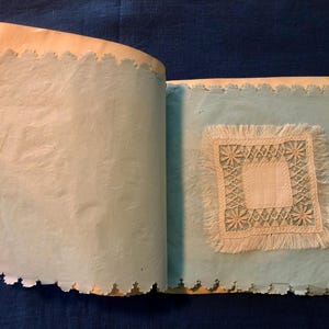 May include: An antique scrapbook with a delicate lace doily adhered to a page. The scrapbook has a light blue paper background and scalloped edges.