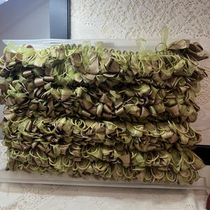 May include: A decorative trim featuring a dense arrangement of looped ribbons in shades of olive green and metallic bronze. The ribbons are layered and attached to a clear plastic header, creating a textured, multi-dimensional fringe effect.