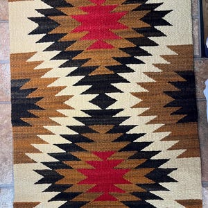 May include: A rectangular woven rug featuring a repeating geometric pattern in cream, brown, black, and red. The design consists of large, symmetrical diamond shapes centered along the length, with horizontal red stripes at the top and bottom edges.