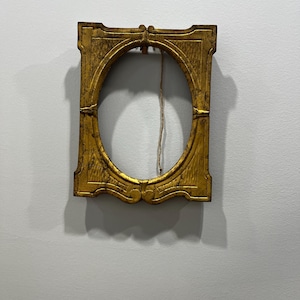 May include: A rectangular gold-colored picture frame with an oval-shaped center opening. The frame features a distressed finish with visible cracks and a weathered texture. A thin, light-colored string hangs vertically from the top center.