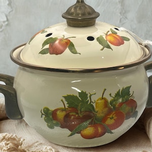 May include: A cream-colored enamel pot with two dark gray handles and a matching lid featuring a decorative fruit pattern of apples, pears, and green leaves. The lid has a dark metallic knob and small steam vents, with a white tag attached.
