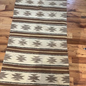 May include: A rectangular woven rug featuring a repeating pattern of horizontal stripes and geometric diamond shapes in shades of cream, dark brown, tan, and black. The textile displays a consistent symmetrical design across its entire surface.