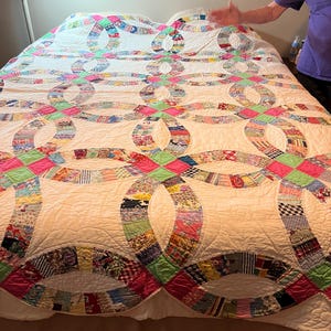 May include: A double wedding ring quilt featuring a white background with interlocking rings composed of various multi-colored fabric scraps. The design includes bright pink and light green solid fabric squares at the intersection points of the rings.