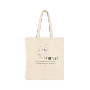 Puede incluir: Bolso tote de color crema con un diseño minimalista. La bolsa presenta un dibujo lineal de una cara con una mariposa y las palabras "LASH NAP". Debajo del texto está la frase: "La única vez que es socialmente aceptable dormir mientras alguien te mira."
