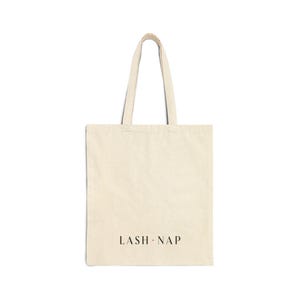 Puede incluir: Un bolso tote de color crema con dos asas. Las palabras "LASH.NAP" están impresas en una fuente marrón oscuro cerca de la parte inferior del bolso. El bolso está hecho de un material similar a la lona y está sobre un fondo blanco.