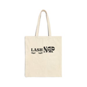 Puede incluir: Un bolso tote de color crema con texto y gráficos negros. El texto dice "LASH NAP" con pestañas estilizadas. El bolso tiene forma rectangular con un asa para llevar. El bolso está hecho de un material similar a la lona.