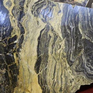 Puede incluir: Una gran losa de piedra natural con un patrón llamativo. La piedra presenta una mezcla de vetas gris oscuro, negras y amarillo dorado, creando una apariencia dinámica y texturizada. El efecto general recuerda al agua que fluye o al arte abstracto.