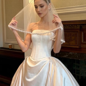 May include: A strapless ivory wedding gown with a full skirt. The bride is wearing a sheer veil with beaded trim. The dress has a fitted bodice and a flowing skirt. The veil is held up by the bride's hands.