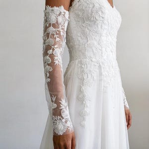 Op de afbeelding: Witte trouwjurk met bloemen kanten details. De jurk heeft een getailleerd lijfje en een zwierige rok. De lange mouwen zijn gemaakt van transparante stof en zijn versierd met ingewikkeld bloemenkant. Een prachtige bruidsjurk voor een speciale gelegenheid.
