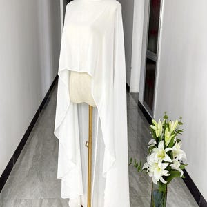 May include: A white, flowing cape-style top on a mannequin. The top has a round neckline and drapes down in a layered design. The mannequin is on a gold stand. A vase of white lilies is in the background.