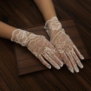 May include: A pair of white lace gloves with floral patterns. The gloves are made of delicate, sheer lace and feature a scalloped edge at the wrist. The gloves are displayed on a dark wooden surface.