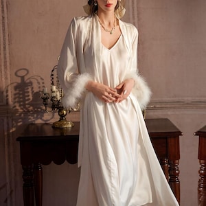 May include: A cream-colored satin slip dress paired with a matching long-sleeve robe. The robe features long sleeves with white feather trim at the cuffs. The slip dress has a V-neckline and a smooth, lustrous finish, creating a coordinated set.