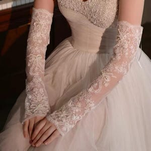 Könnte beinhalten: Ein trägerloses elfenbeinfarbenes Brautkleid mit einem bestickten Oberteil und einem vollen Tüllrock. Die Braut trägt lange, zarte elfenbeinfarbene Spitzenhandschuhe mit floralen Mustern und Perlenakzenten. Die Handschuhe reichen vom Handgelenk bis zum Oberarm.