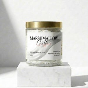 Puede incluir: Un tarro de vidrio transparente con tapa dorada, lleno de manteca corporal batida blanca. La etiqueta dice "Marshmallow Chiffon, Whipped Body Butter, 8 oz." El tarro está sobre un bloque blanco.
