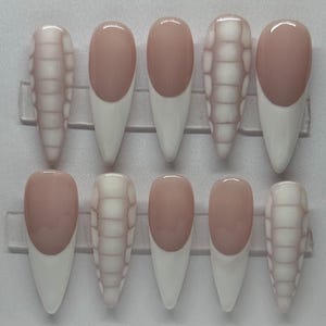 Puede incluir: Un conjunto de diez uñas artificiales de varias formas. Las uñas presentan puntas blancas, una base de color nude y un diseño texturizado en algunas. Están dispuestas en un soporte de plástico transparente.