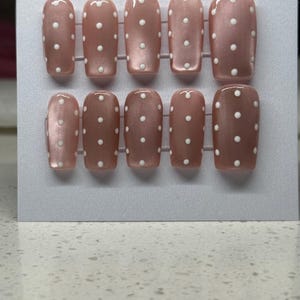 May include: Set of ten artificial nails in a light brown shade, each adorned with small white polka dots. The nails are arranged on a white card, showcasing a classic and feminine design. The nails have a glossy finish.