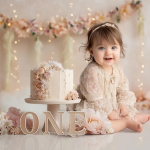 Könnte beinhalten: Eine Geburtstagsszene mit einem mit Blumen geschmückten Kuchen, einem Holzschild 'ONE' und einem lächelnden Kind. Der Kuchen steht auf einem Ständer, mit Blumendekorationen und sanfter Beleuchtung im Hintergrund. Der Kuchen ist etwa 15 cm hoch.