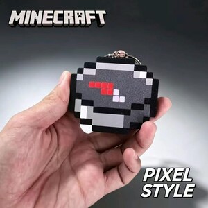 Puede incluir: Un llavero con temática de Minecraft que presenta un diseño de brújula circular pixelada. La esfera es principalmente gris y negra con un indicador de aguja pixelado en rojo y blanco. Incluye una anilla metálica plateada arriba.