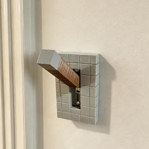 May include: A light switch cover plate featuring a grid pattern in a matte light grey finish. The toggle switch is extended with a rectangular wooden handle that has a light grey tip, creating a distinct contrast against the grey tiled plate.