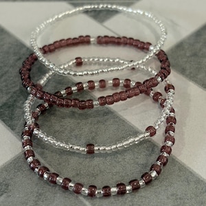 May include: Four beaded bracelets. Two bracelets are silver with clear beads, and two are silver with dark purple beads. The bracelets are stacked on top of each other. The background is a gray and white checkered pattern.