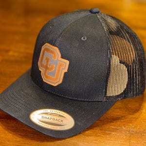 Puede incluir: Gorra de camionero negra con parte trasera de malla y visera curva. La gorra presenta un parche de cuero con un logotipo CU naranja y marrón. Tiene cierre a presión y un pequeño botón en la parte superior.