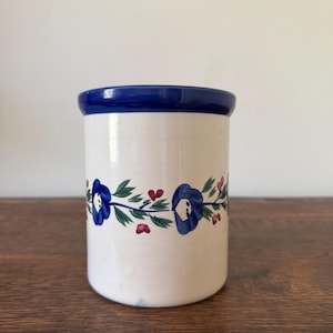 May include: A cylindrical ceramic crock with a cream-colored base and a deep blue rim. The exterior features a hand-painted horizontal band of blue flowers, green leaves, and small red berries encircling the center of the vessel.