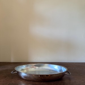 May include: A round, shallow silver-toned metal serving dish with a polished, reflective finish. The dish features two small, decorative handles on opposite sides, each with a textured, scalloped edge design. The interior is smooth and concave.